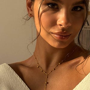 Op de afbeelding: Een goudkleurige ketting met een fijne ketting en kleine bolvormige kralen. De ketting heeft een Y-vormige hanger. Het model draagt een witte trui en gouden oorbellen. De foto is goed belicht.