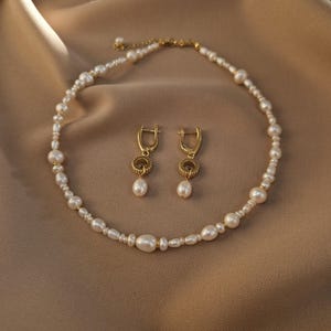 May include: A pearl necklace and matching earrings set. The necklace and earrings feature white pearls and gold-coloured metal accents. The earrings have a circular design with a dangling pearl. The jewellery set is displayed on a tan fabric.