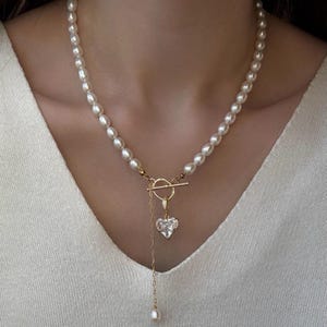 May include: A necklace featuring a strand of white oval pearls with a gold-toned toggle clasp. A gold-toned chain hangs from the clasp, ending in a single small pearl, while a heart-shaped clear crystal pendant is attached to the circular toggle ring.