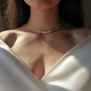 以下が含まれることがあります： 鎖骨に沿って中央に5つの小さな丸い白い真珠が並んだ、繊細なゴールドチェーンのネックレス。ネックレスは滑らかなオフホワイトの生地の衣服の上に着用されており、金属製のゴールドチェーンと光沢のある白い真珠のコントラストが際立っています。