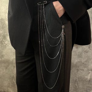 May include: Silver chain accessory for pants. The accessory features multiple strands of delicate chains, some with small beads, attached to safety pins. The chains hang vertically, creating a layered effect. The accessory is attached to a black suit.