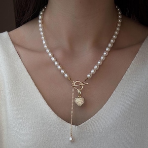 May include: A necklace featuring a strand of white oval pearls connected to a gold-toned toggle clasp. A gold-toned chain hangs from the clasp, ending in a small pearl, alongside a gold-toned heart-shaped pendant covered in small clear stones.