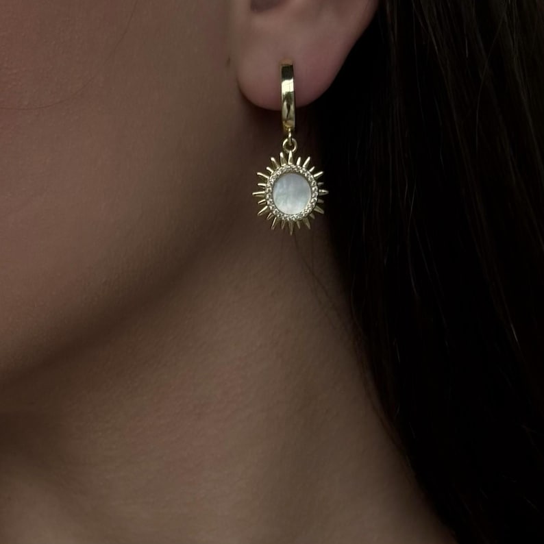 Peut inclure: Une boucle d'oreille cr&eacute;ole en or avec un pendentif en forme de soleil rayonnant, dot&eacute; d'une incrustation centrale circulaire en nacre blanche. Le motif solaire pr&eacute;sente des rayons m&eacute;talliques entourant la pierre iris&eacute;e.