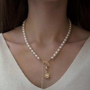 May include: A pearl necklace with a gold clasp and a sun pendant. The necklace features a strand of white pearls and a gold-colored sunburst charm with small, sparkling accents. The clasp is a gold bar and ring design.