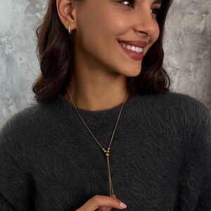A woman wears a gold Delicate Y Necklace with small beads and an adjustable slider. The Dainty Jewelry Set includes matching Small Hoop Earrings.