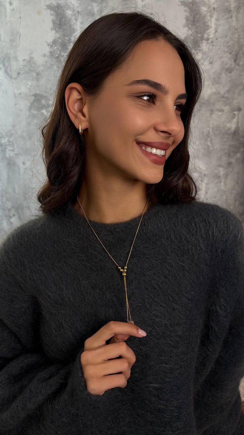 A woman wears a gold Delicate Y Necklace with small beads and an adjustable slider. The Dainty Jewelry Set includes matching Small Hoop Earrings.