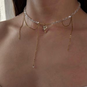 Könnte beinhalten: Eine zarte Halskette mit einem durchsichtigen Kristall-Choker mit goldfarbenen Kettenakzenten. Die Halskette hat einen kleinen goldfarbenen Blumenanhänger und baumelnde Ketten. Die Halskette ist zum Tragen um den Hals konzipiert.