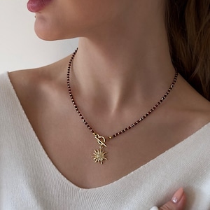 May include: Gold-tone necklace with a sunburst pendant and matching stud earrings. The necklace features a strand of small, dark red beads. The pendant and earrings have a detailed, textured design.