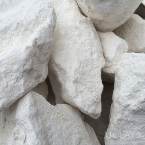 May include: Close-up of a pile of white clay lumps. The clay is rough and textured. The website address "UCLAYS.COM" is visible in the bottom right corner of the image.
