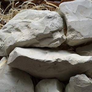 Puede incluir: Primer plano de varios trozos grandes de arcilla gris claro. Las piezas de arcilla tienen superficies rugosas e irregulares y están dispuestas en una cesta de mimbre con paja. La arcilla parece ser un producto natural.