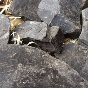 Puede incluir: Primer plano de varias piezas de nibs de cacao crudos de color gris oscuro y forma irregular. Los nibs están apilados en una cesta.