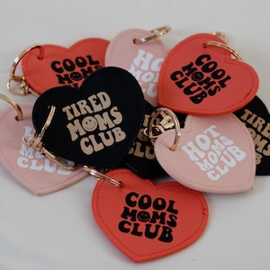 Może przedstawiać: Zbiór breloków w kształcie serca z różnymi napisami, w tym "Cool Moms Club", "Tired Moms Club" i "Hot Moms Club". Każdy brelok zawiera grafikę uśmiechniętej twarzy i jest wykonany ze skóry.