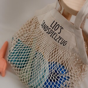 Op de afbeelding: Een beige netzak met een canvas bovenkant. De canvas bovenkant heeft de tekst "Lio's Sandspielzeug" geborduurd. De tas is gevuld met blauwe en groene plastic zand speelgoed.