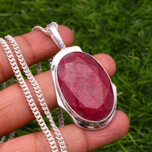 May include: A silver chain necklace with a pendant featuring a large, oval-shaped ruby gemstone. The ruby is a deep red colour and has a faceted cut.