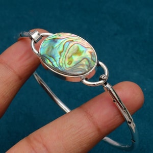 May include: A silver bangle bracelet featuring an oval abalone shell centerpiece. The shell displays iridescent colors of green, blue, and yellow. The bracelet has a simple, elegant design with a hinged closure.