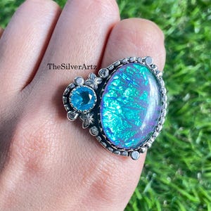 Op de afbeelding: Een zilveren ring met een grote ovale iriserende blauwe en paarse steen naast een kleinere ronde blauwe steen. De metalen zetting heeft decoratieve bloemachtige accenten en de tekst TheSilverArtz is zichtbaar op het oppervlak van de ring.