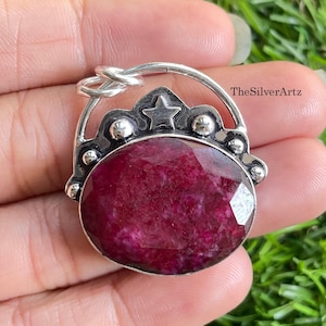 May include: A silver pendant featuring a large, oval-shaped, faceted deep pink gemstone. The silver setting includes a decorative arched top with a small star cutout, three small silver spheres, and a circular bail. The text TheSilverArtz is visible.