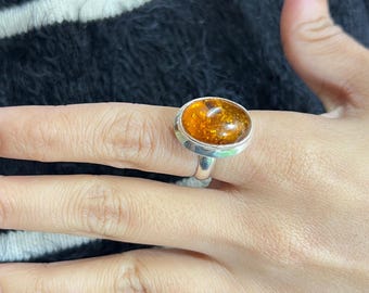 May include: A silver ring featuring a large, oval-shaped, polished amber gemstone with a warm, translucent orange-brown hue. The stone is set in a smooth, plain silver bezel mount that sits atop a simple, polished silver band.