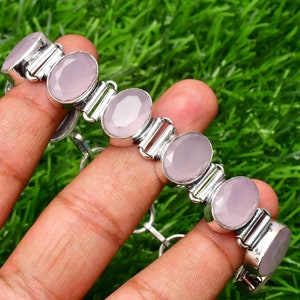 Puede incluir: Una pulsera de plata con piedras preciosas ovaladas de color rosa pálido. Las piedras están engastadas en un marco de metal plateado y unidas por eslabones plateados. La pulsera se muestra sobre un fondo verde.