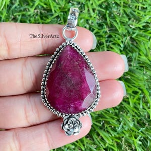 May include: A teardrop-shaped pendant featuring a faceted deep pink gemstone set in a silver frame with a beaded edge and a small floral accent at the bottom. The pendant includes a silver bail at the top and the text TheSilverArtz is visible.