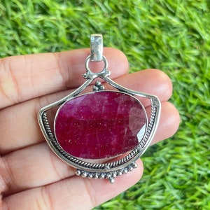 May include: A silver pendant featuring a large, oval-cut ruby gemstone. The ruby is a deep, vibrant red color and is set within a decorative silver frame. The pendant has a bail for attaching to a chain.