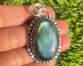 Puede incluir: Un colgante de plata con una gran piedra ovalada de labradorita con reflejos azules y verdes iridiscentes. La piedra está engastada en un engaste de plata con un borde decorativo de pequeñas cuentas de plata.