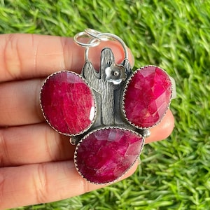 Könnte beinhalten: Ein silberner Anhänger mit einem stilisierten Kaktus-Design und drei roten Rubin-Steinen. Der Anhänger hat oben eine Schlaufe zum Anbringen einer Kette.