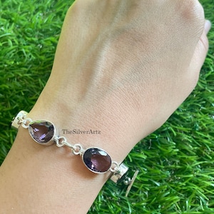 May include: A silver link bracelet featuring alternating teardrop and oval-shaped purple amethyst gemstones. The metal links connect each stone, and the text TheSilverArtz is visible near the center of the bracelet, which is worn on a wrist.