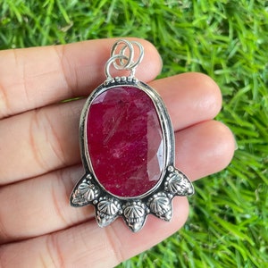 May include: A silver pendant with a large, oval-shaped, red ruby gemstone. The pendant has a detailed, ornate design with five points at the bottom.