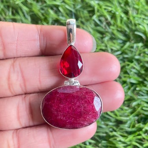 以下が含まれることがあります： 赤いルビーの石が2つ付いたシルバーのペンダント。上の石は涙型、下の石は楕円形。ペンダントは手に置かれている。