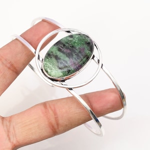 May include: A silver bangle bracelet with a green and pink oval gemstone set in a circular design.
