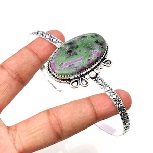 May include: A silver-toned cuff bracelet featuring an oval-shaped gemstone with green and pink hues. The bracelet has a decorative floral design around the gemstone and a geometric pattern on the band.