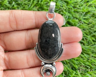 May include: A black oval pendant with a silver bail and decorative accents. The pendant features a dark, speckled stone set in a silver frame. The bottom of the pendant has a crescent moon design with a small floral detail.