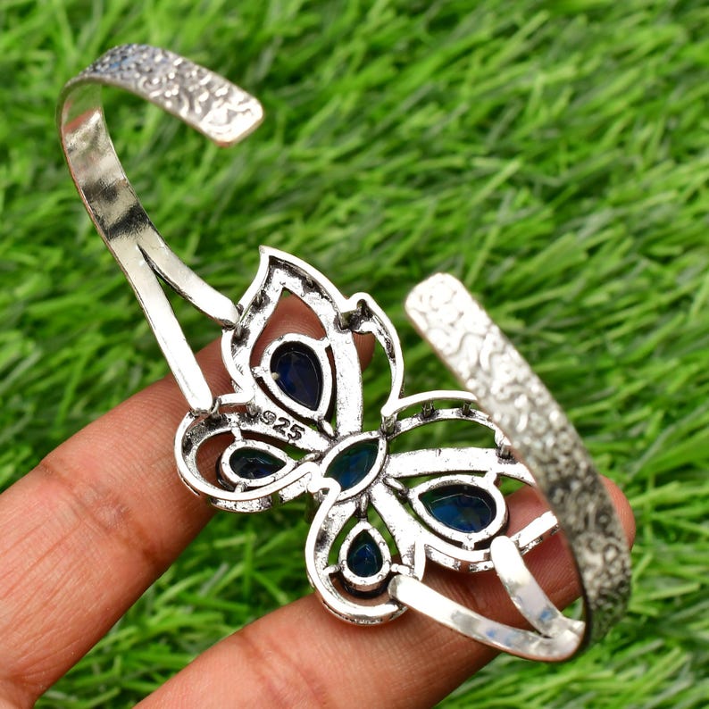 May include: Silver butterfly bangle bracelet with blue gemstone accents. The bracelet is open and has a textured finish. The back of the bracelet is stamped with "925".