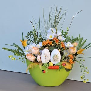 Könnte beinhalten: Eine leuchtend grüne Schale, die mit einem Osterblumenarrangement überquillt. Das Arrangement umfasst weiße, orangefarbene und pfirsichfarbene Blumen, zwei dekorierte Ostereier und dekorative Karotten. Ein festlicher Frühlings-Mittelpunkt.