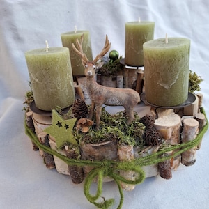 Könnte beinhalten: Ein rustikaler Adventskranz mit vier grünen Kerzen, einer kleinen Hirschfigur, Tannenzapfen und einem grünen Stern. Der Kranz besteht aus Birkenholzstämmen und Moos, mit einer grünen Ranke um die Basis. Eine festliche Weihnachtsdekoration.