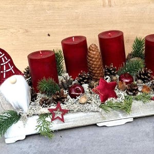Può includere: Un centrotavola natalizio festivo con quattro candele rosse, pigne e decorazioni a stella rossa. Uno gnomo con la barba bianca con un cappello rosso e bianco è a sinistra. La composizione è in un vassoio di legno bianco.