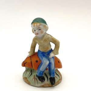 May include: A porcelain figurine of a boy wearing a green hat and blue pants sitting on a red and orange ladybug.