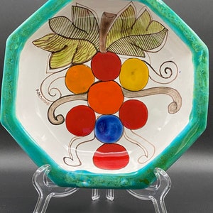May include: A white ceramic octagonal serving bowl with a turquoise rim. The bowl features a hand-painted design of a bunch of grapes with green leaves, red, orange, and yellow grapes, and a blue grape in the center. The bowl is signed "R.BRESCIONE".