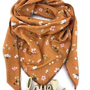 May include: A rust-colored scarf with a floral pattern of white and pink flowers and green leaves. The scarf has a triangular shape and is decorated with a wooden "love" sign.
