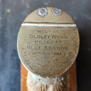 May include: A brass bottle stopper with a round top and a fur-covered bottom. The top has an inscription that reads "MEET AT SLOLEY WOOD KILLED AT BLUE ANCHOR 29th JULY 1823 GIVEN TO WASBROUGH BY THE MASTER."