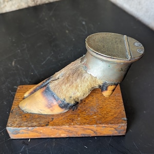 May include: A vintage, taxidermied animal foot inkwell with a brass lid. The lid has the inscription "Solid Wood Black Filled Inkwell". The foot is mounted on a wooden base.