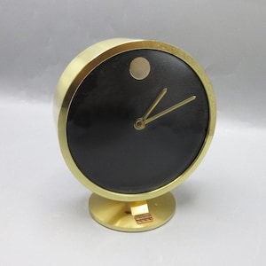 May include: A gold-toned, round clock with a black face and gold hands. The clock has a small, gold dot at the 12 o'clock position and sits on a matching gold base. The clock's design is minimalist and elegant.