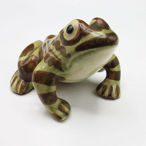 May include: A ceramic frog figurine featuring a glossy finish with a light green base color and dark brown horizontal stripes across its body and legs. The frog has large, prominent eyes with dark pupils and a smooth, rounded head shape.