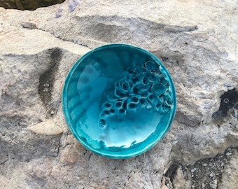 May include: A teal blue ceramic dish with a raised floral design. The dish is round and has a glossy finish.