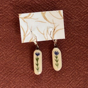 May include: A pair of dangle earrings featuring elongated, cream-colored ceramic pendants with a hand-painted blue flower and green stem design. The pendants hang from silver-toned metal ear wires attached to a white display card with brown lines.