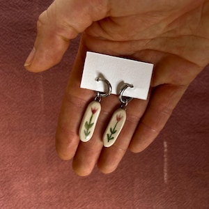Peut inclure: Une paire de boucles d'oreilles créoles en argent avec des pendentifs ovales en céramique blanche. Chaque pendentif est orné d'une illustration peinte à la main d'une tulipe rouge avec des feuilles et une tige vertes, suspendue à un anneau en argent.