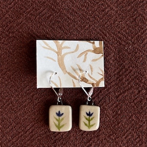 Puede incluir: Un par de pendientes colgantes con dijes rectangulares de cerámica color hueso, decorados con un diseño pintado a mano de una flor azul y un tallo verde. Los dijes cuelgan de ganchos de metal plateado y están sujetos a una tarjeta blanca.