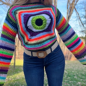 May include: A colorful crocheted sweater with a large eye design in the center. The sweater has long sleeves and is made with a variety of colors, including red, orange, yellow, green, blue, purple, and black.