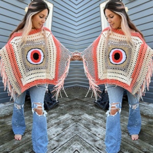 Puede incluir: Un poncho de crochet con un gran diseño de ojo en el centro. El poncho está hecho con una combinación de hilo beige, coral y blanco y tiene flecos alrededor de los bordes.
